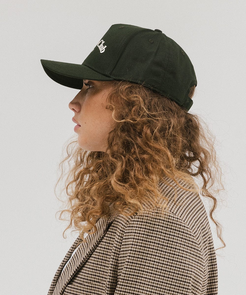 Woman wearing a dark green cap with 'Vacation Club' text and a coat, facing sideways against a plain background  #color_dark green