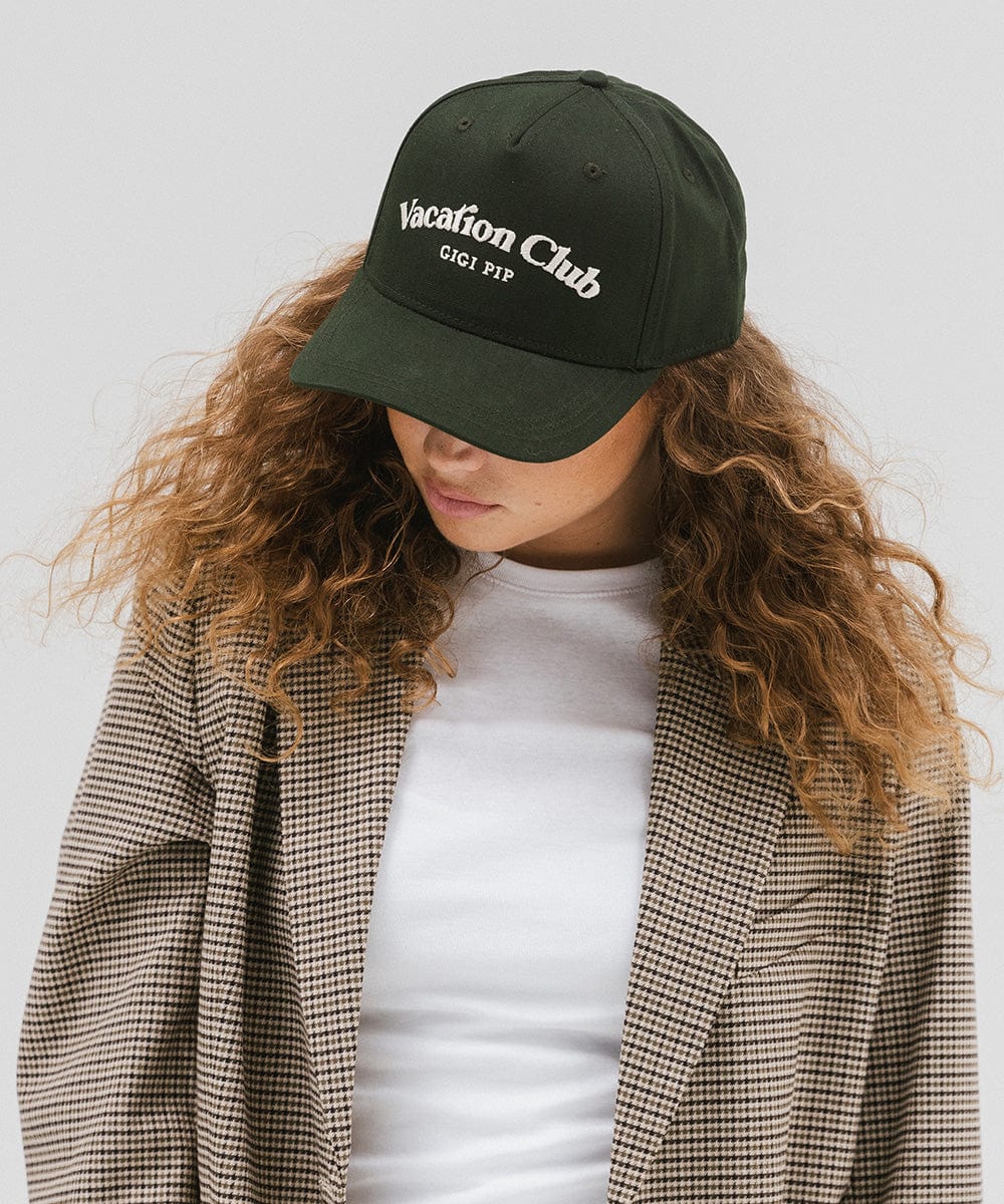 Woman wearing a dark green cap with 'Vacation Club' text and a white top with a coat against a plain background  #color_dark green