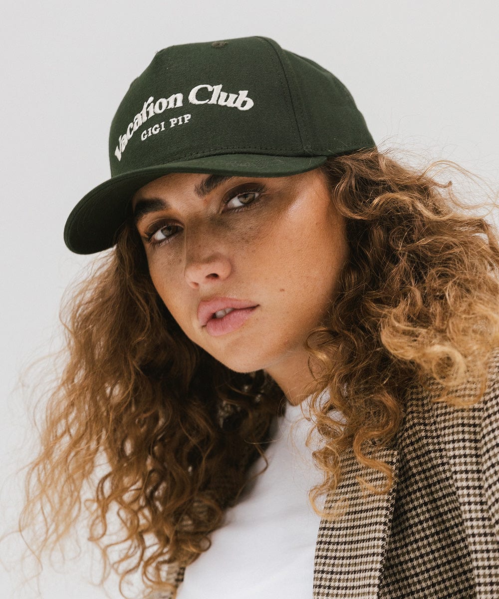 Woman wearing a dark green cap with 'Vacation Club' text and a white top against a plain background  #color_dark green