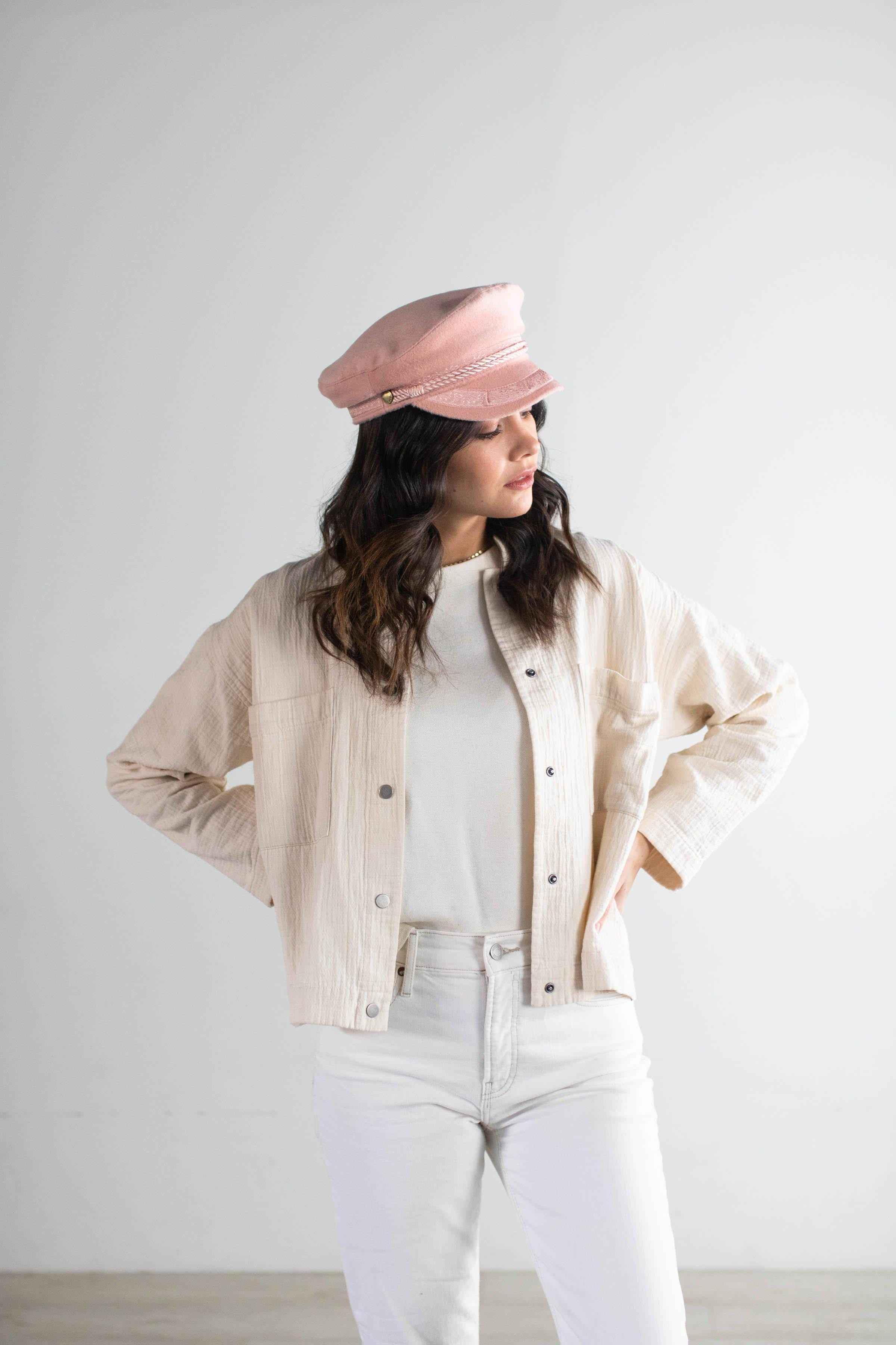 Woman wearing a blush colored lieutenant cap, a white t-shirt with a cream jacket and white pants, posing sideways against a plain background #color_blush