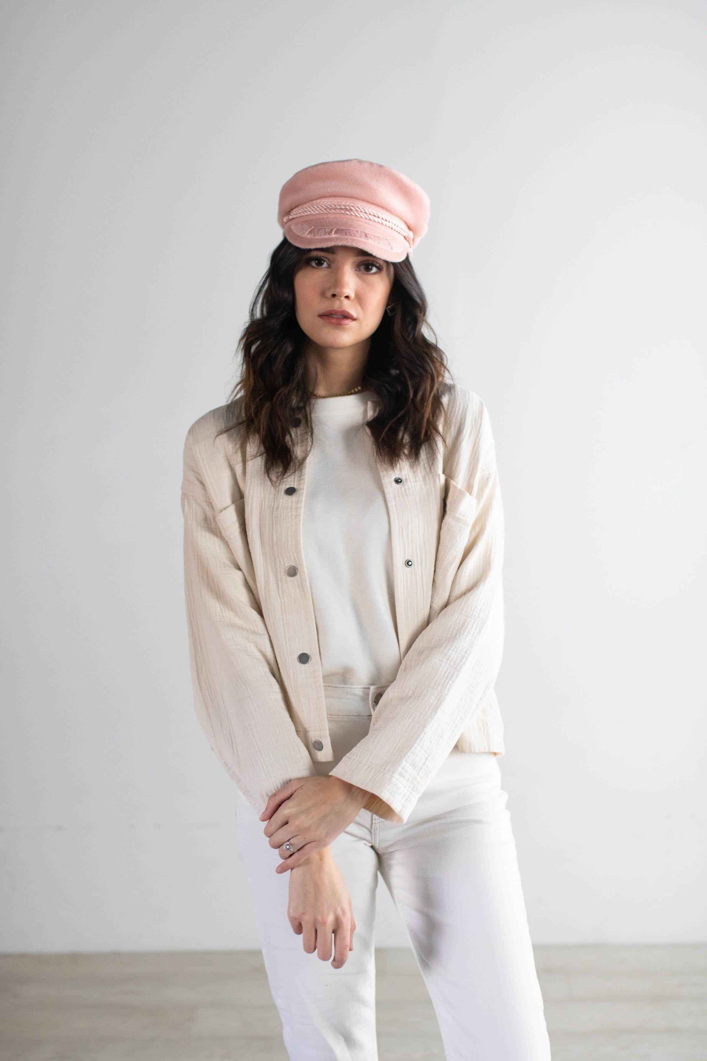 Woman wearing a blush colored lieutenant cap, a white t-shirt with a cream jacket and white pants against a plain background #color_blush