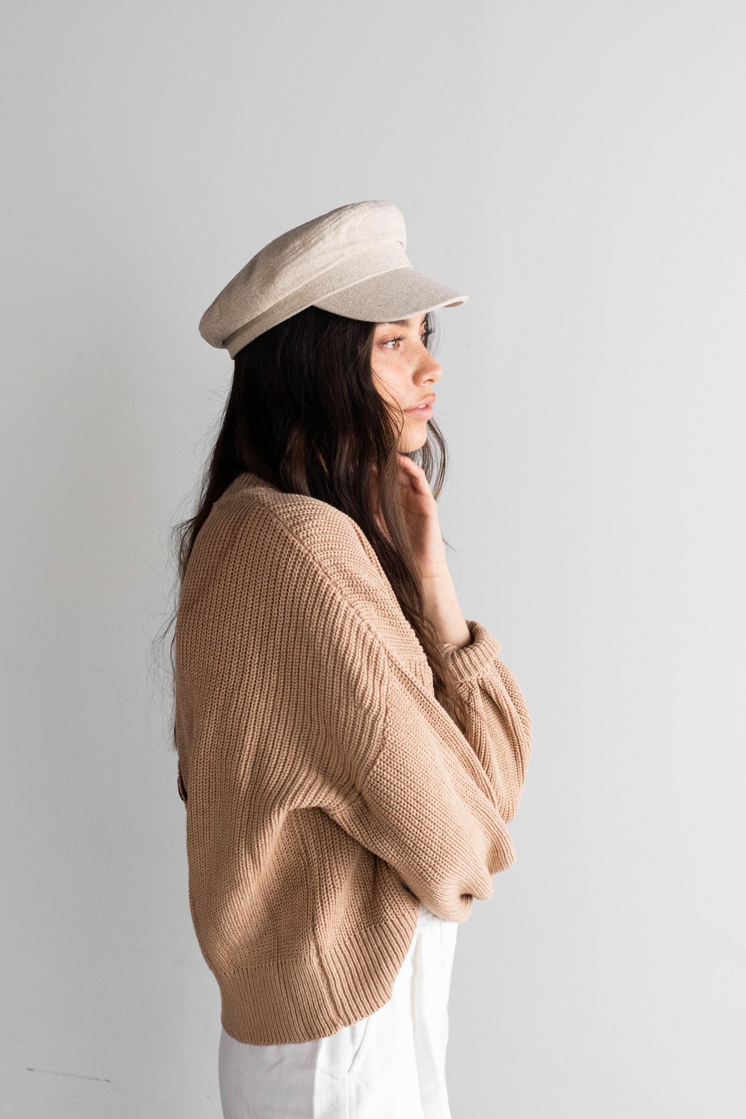 Woman wearing a linen colored cap, a brown sweater and white pants, facing sideways against a plain background #color_linen