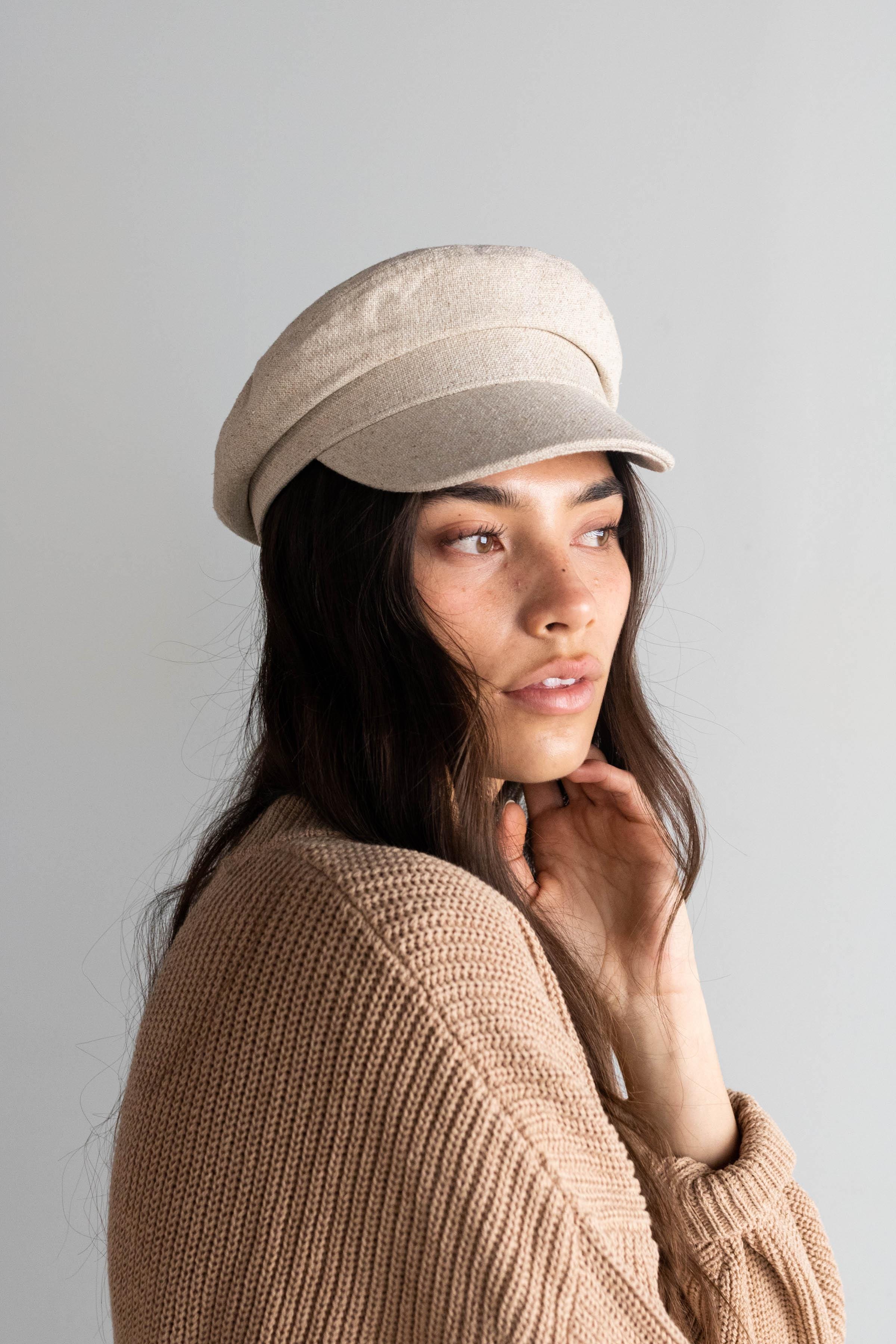 Woman wearing a linen colored cap and a brown sweater, posing sideways against a plain background #color_linen
