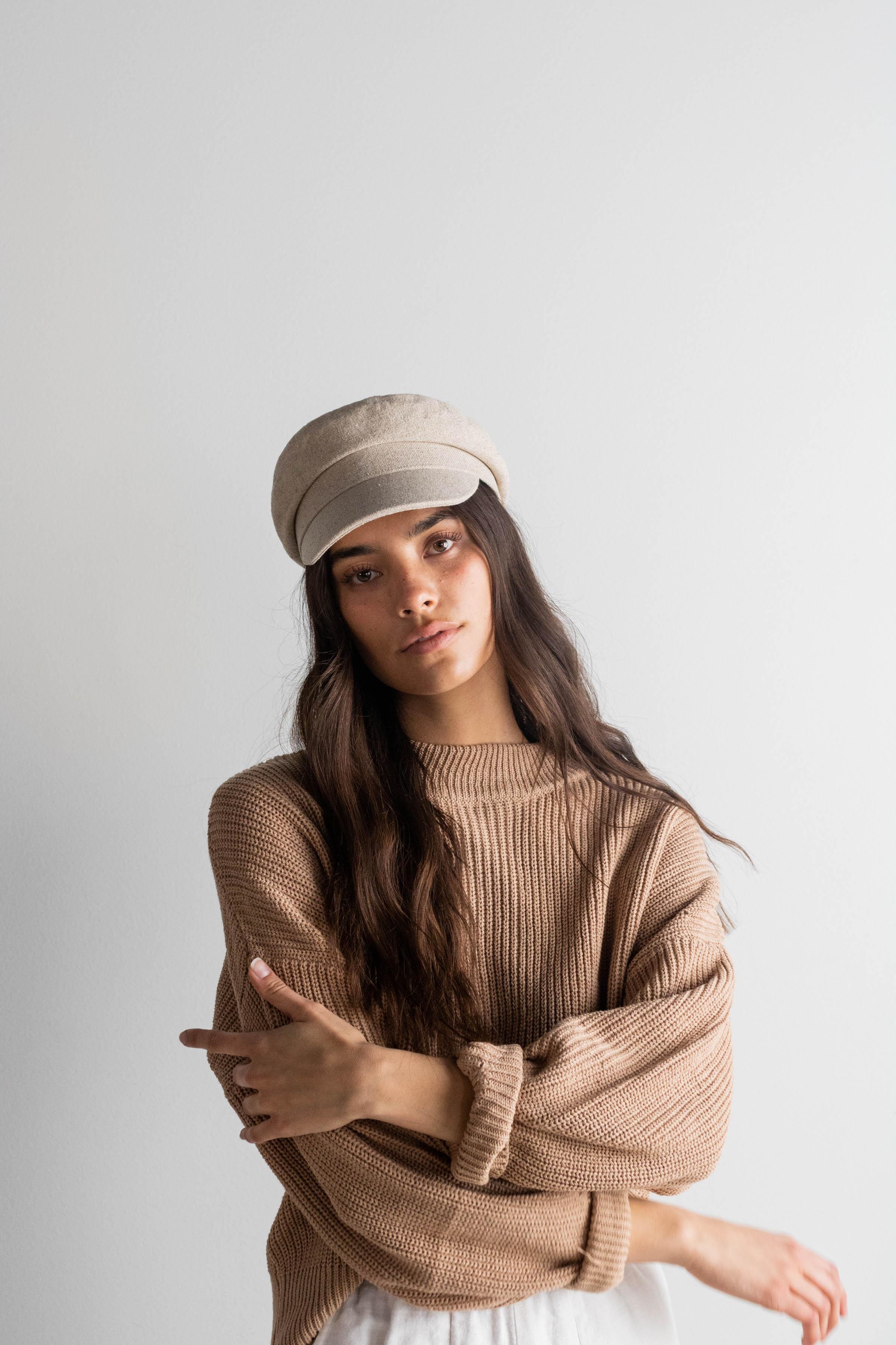 Woman wearing a linen colored cap and a brown sweater against a plain background #color_linen