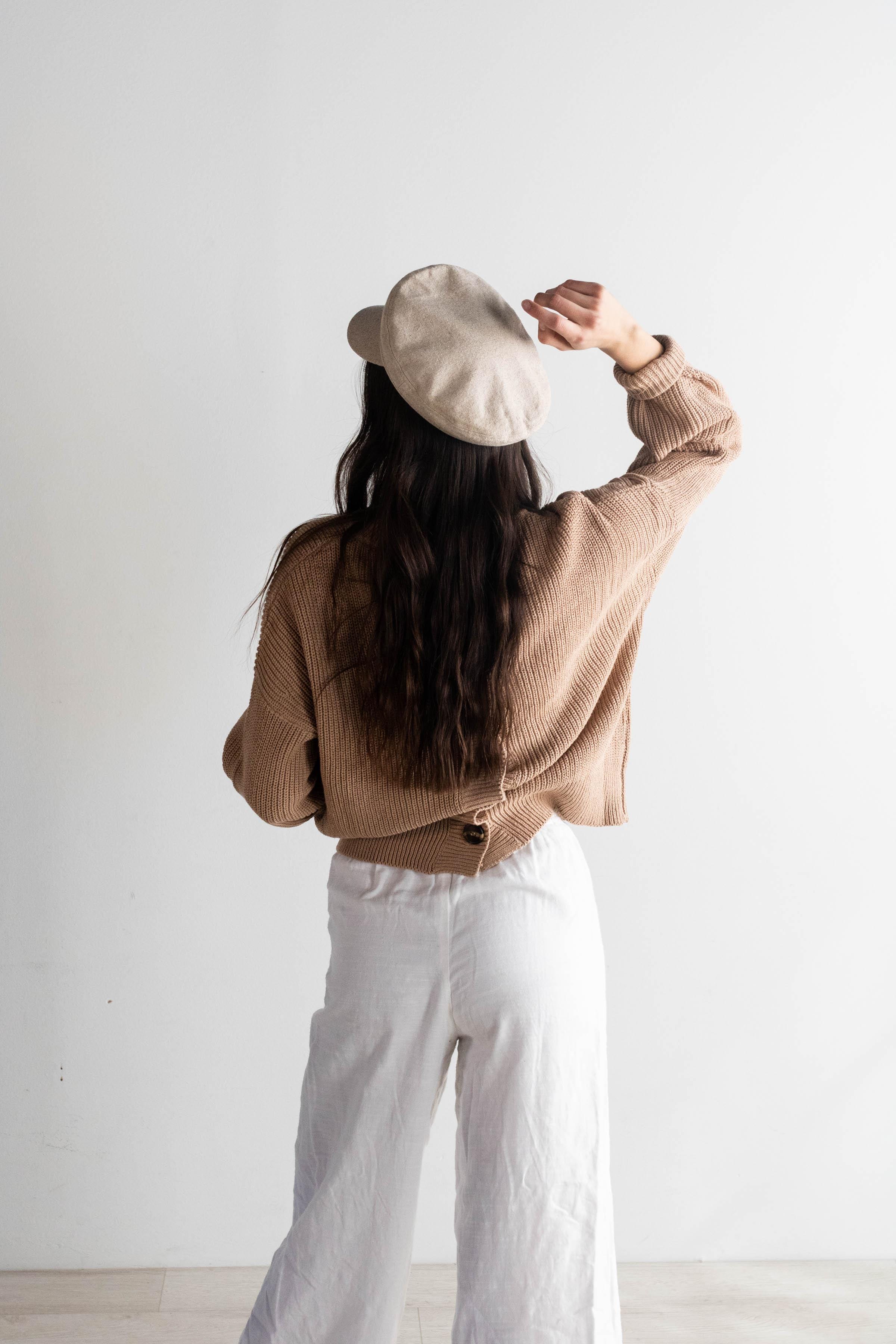 Woman wearing a linen colored cap,  a brown sweater and white pants, posing against a plain background #color_linen
