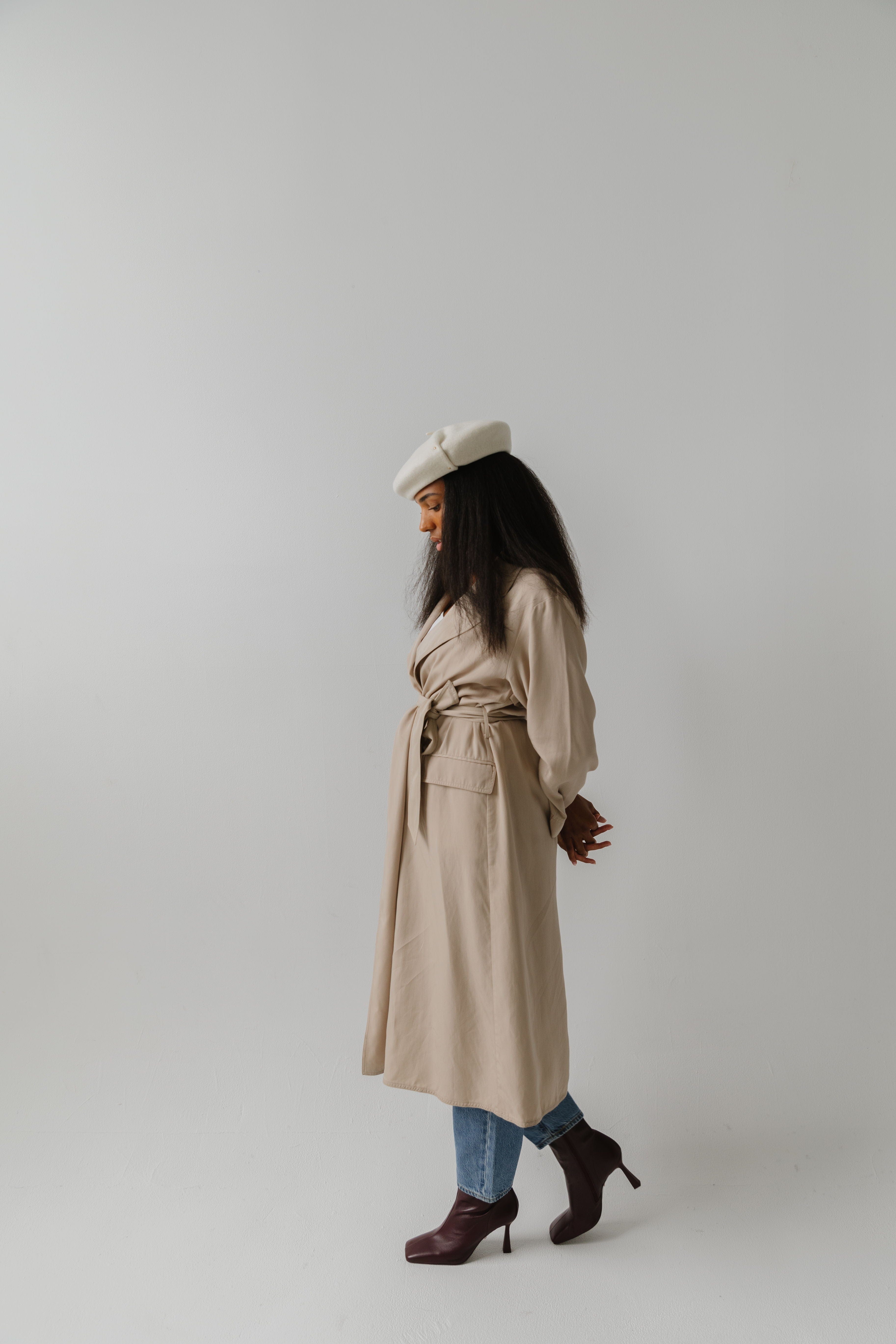 Woman wearing an off white beret, a beige coat, jeans and brown boots, posing against a plain background  #color_off white