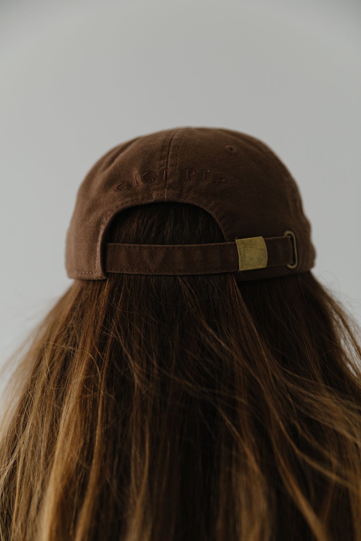 Woman wearing a chocolate colored ball cap facing behind against a plain background #color_chocolate