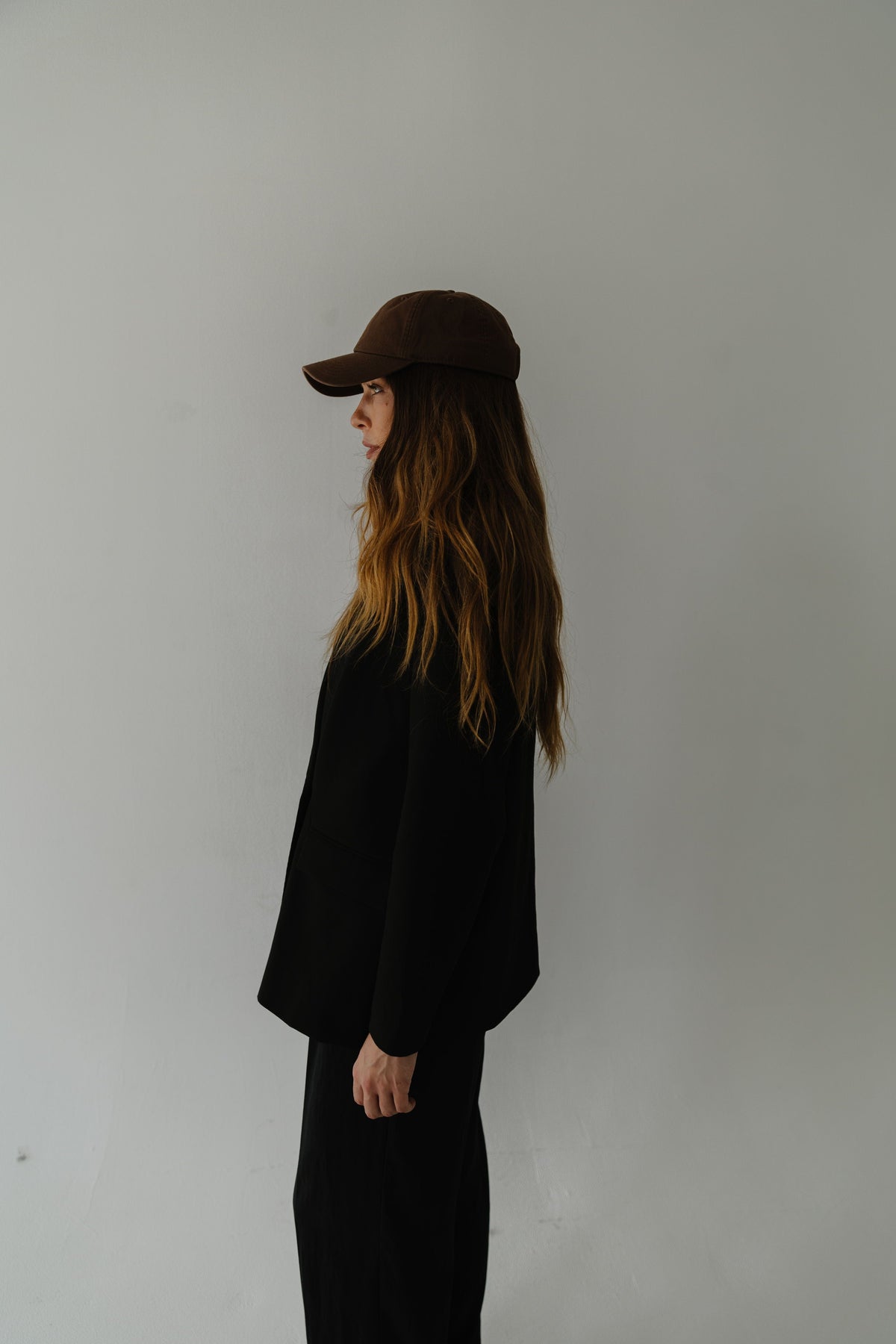 Woman wearing a chocolate colored ball cap, a black shirt and black pants, facing sideways against a plain background #color_chocolate